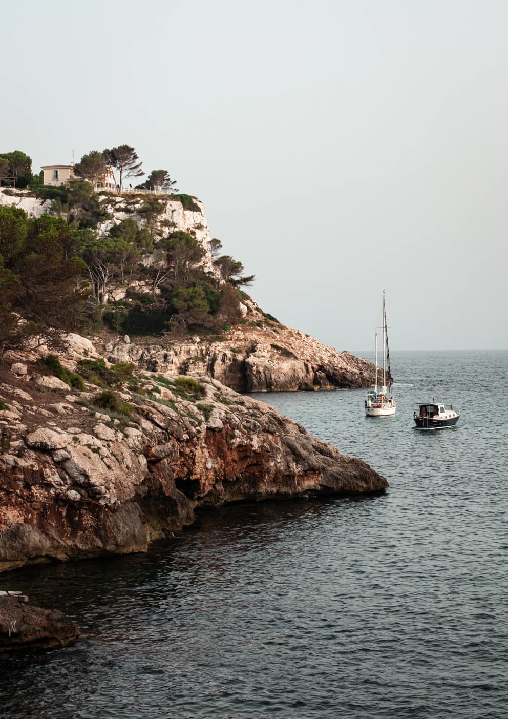 Vela: a sailboat and a small motorboat anchored below a pine-covered headland with a villa on top