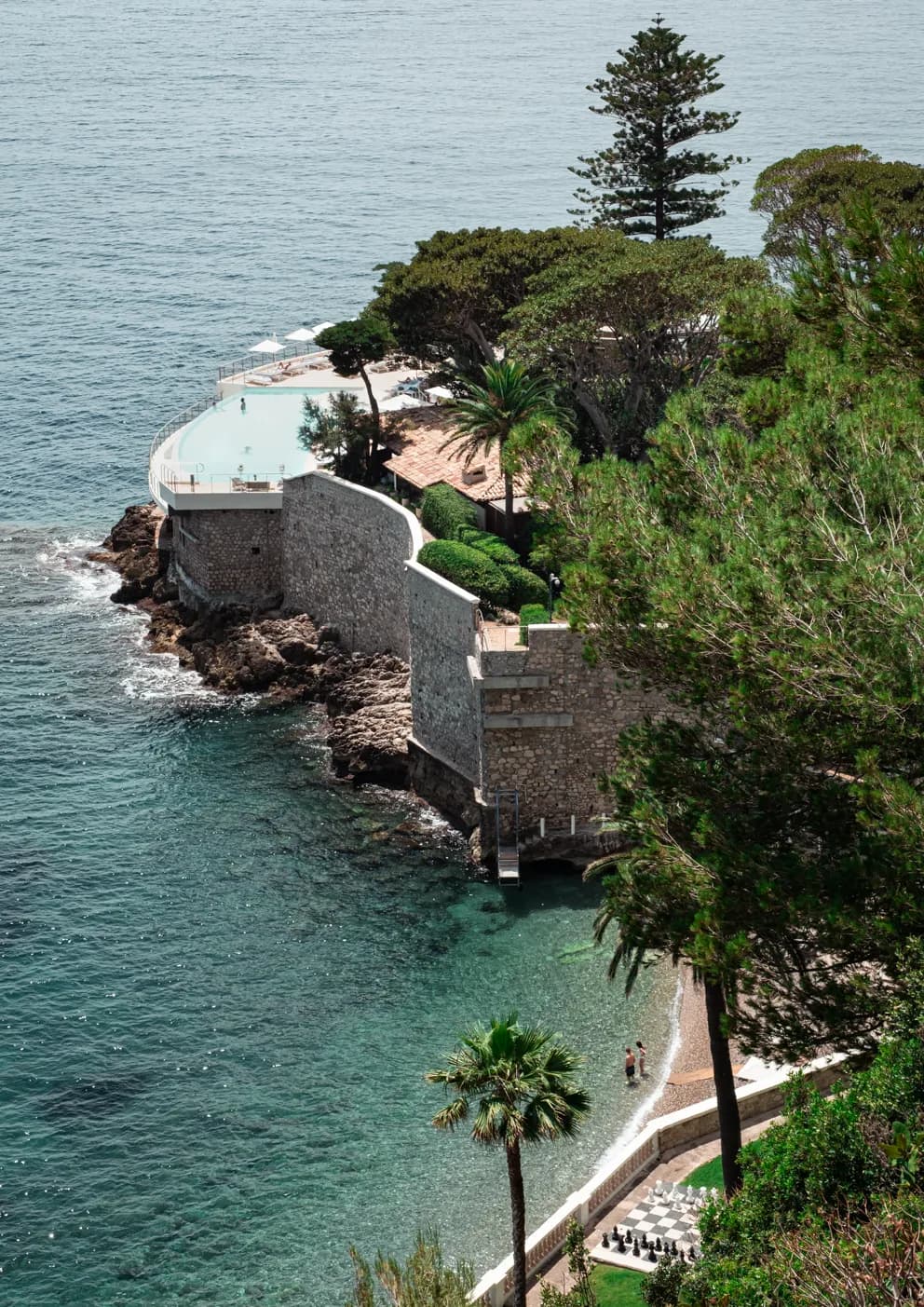 Piscine: a pool perched on stone walls above the sea, with pines, palms and a small beach below
